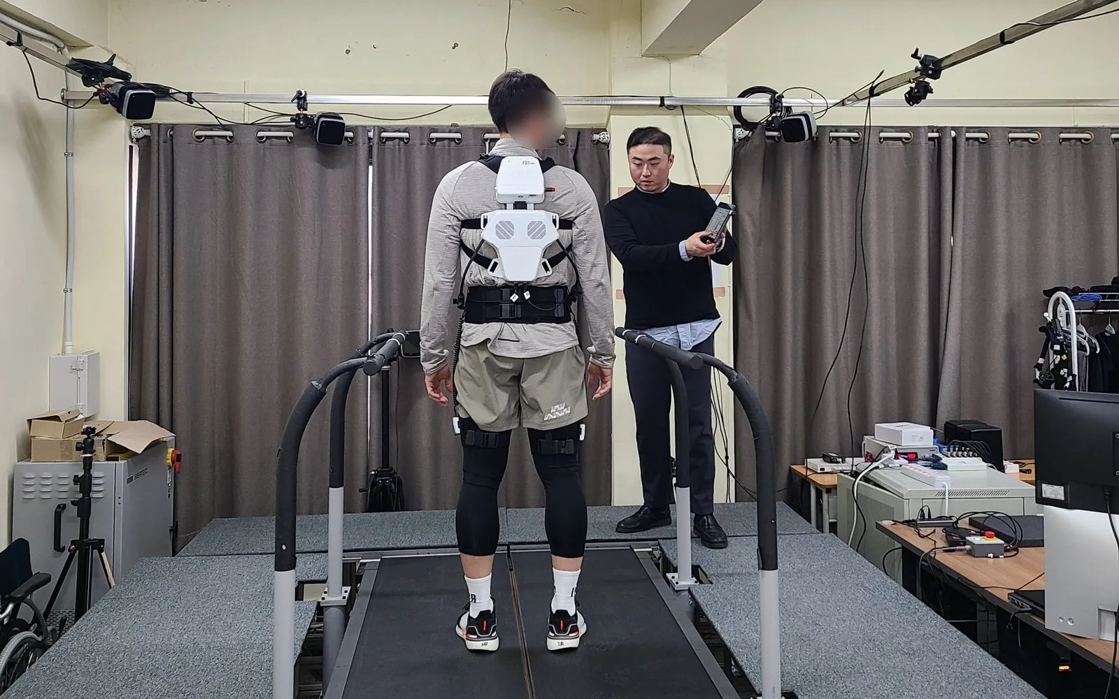 Un hombre camina en una cinta con el exotraje H-Medi de Hurotics mientras un técnico ajusta la asistencia con una tableta, en un laboratorio de biomecánica
