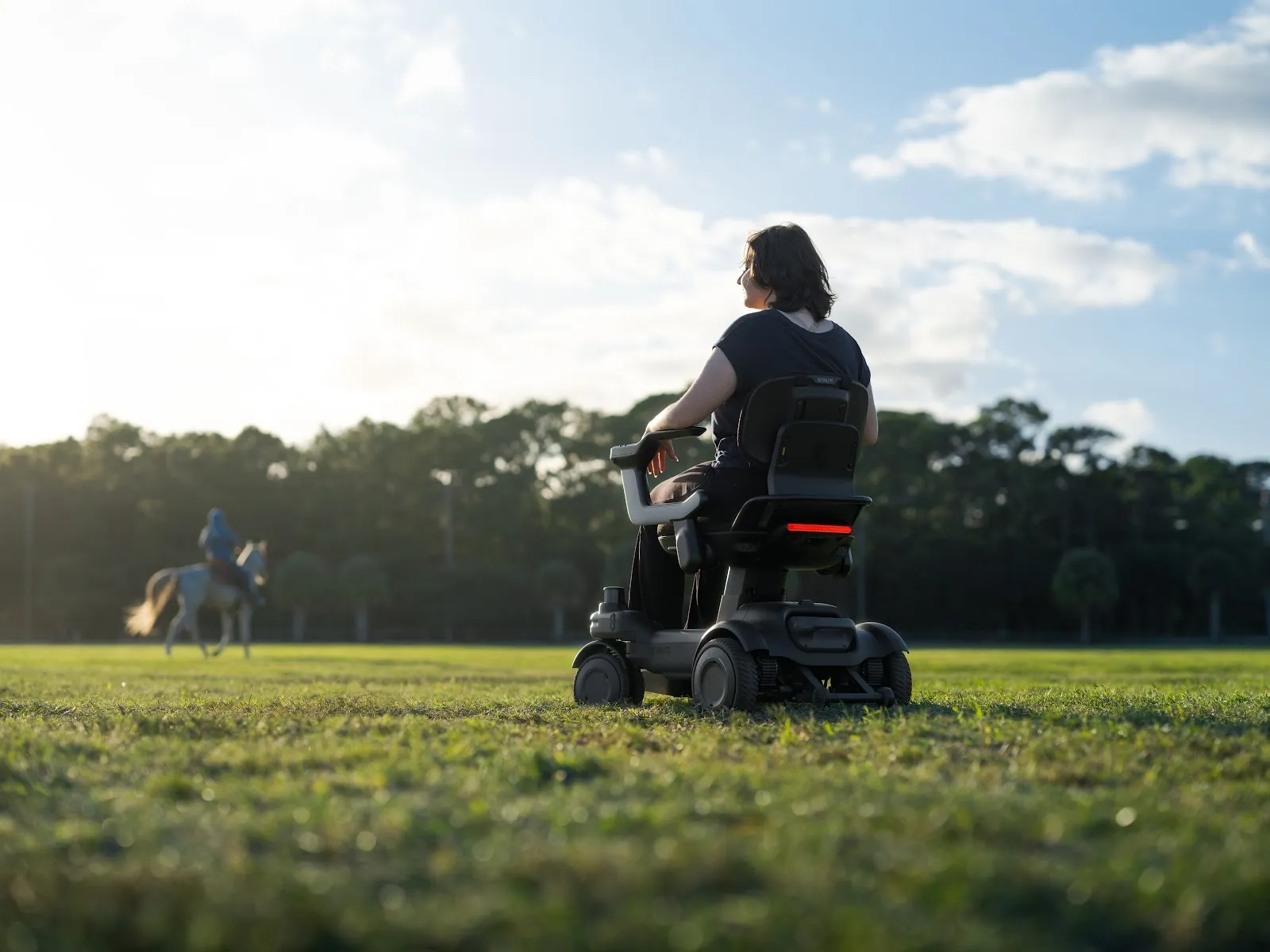 Una mujer conduce una silla eléctrica Strutt ev¹ por una gran pradera de césped al atardecer, vista desde detrás