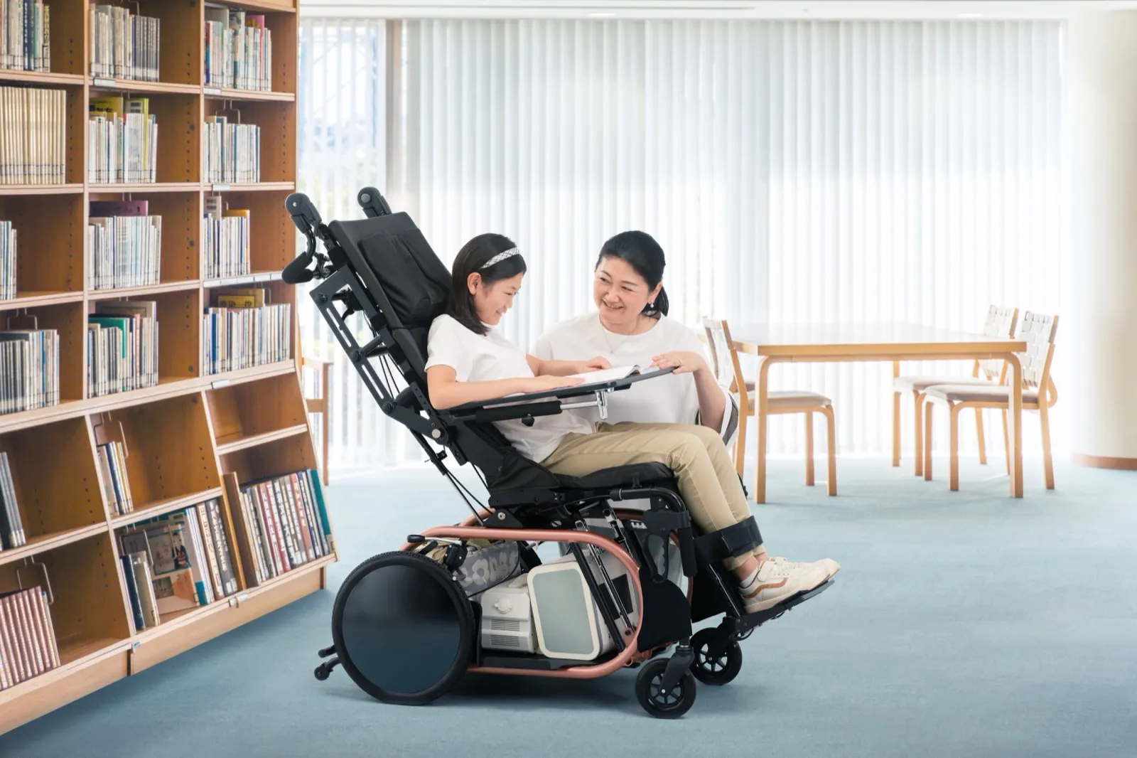 Una niña sentada en la silla de ruedas basculante Toyota Haluto lee un libro junto a su madre en una biblioteca